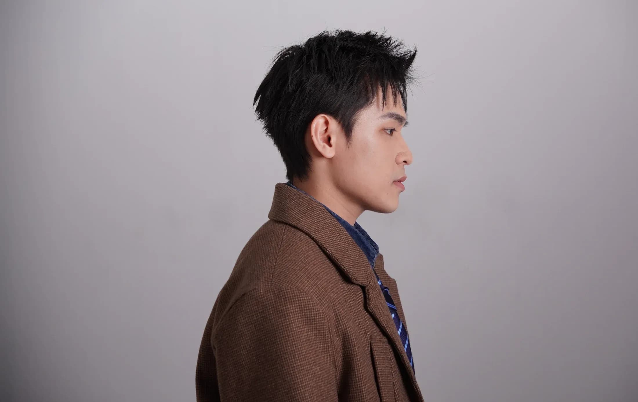 KIDEN in profile wearing a brown jacket against a grey studio background